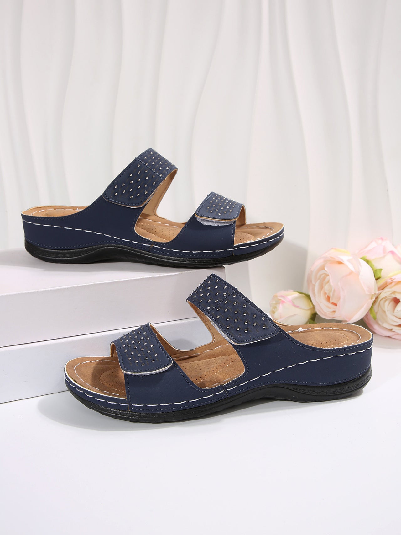 Pair of navy sandals with brown soles on a white surface with flowers in the background